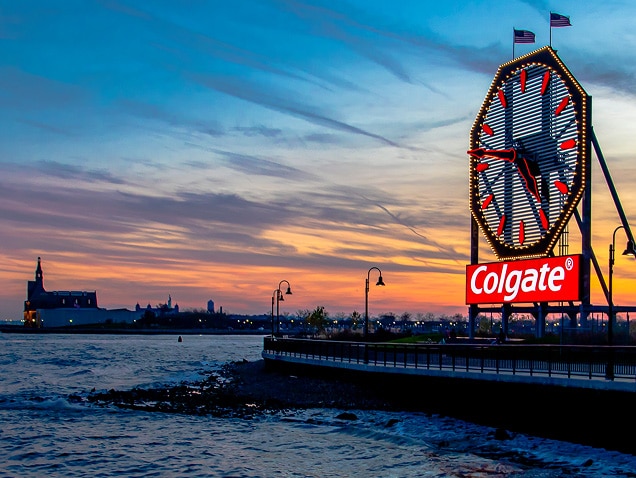orilla del mar, logo de Colgate al fondo bajo un gran reloj con manecillas rojas